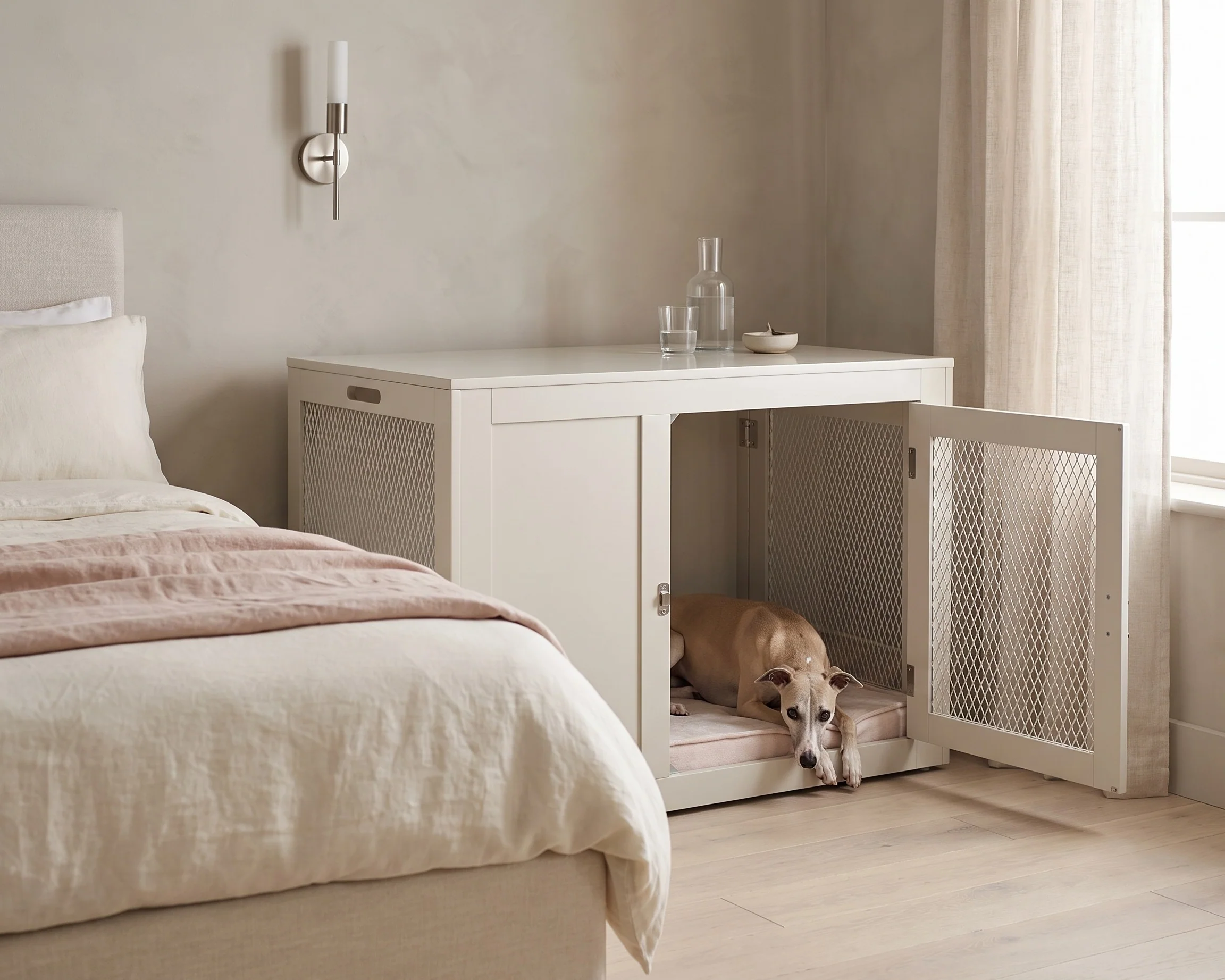A wire dog crate set up beside a bed with a light cover over the back and sides, a simple crate mat inside, and a water bowl clipped to the crate